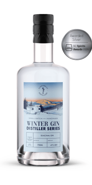 A clear glass bottle with clear liquid in it and a label on the front. A blue label with an illustration of a caravan in front of a hill landscape, titled Winter Gin.