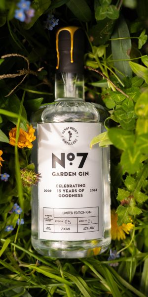 A clear glass bottle of gin with a label on the front titled "No.7 Garden Gin". The bottle is placed in a field pf green grasses and flowers.