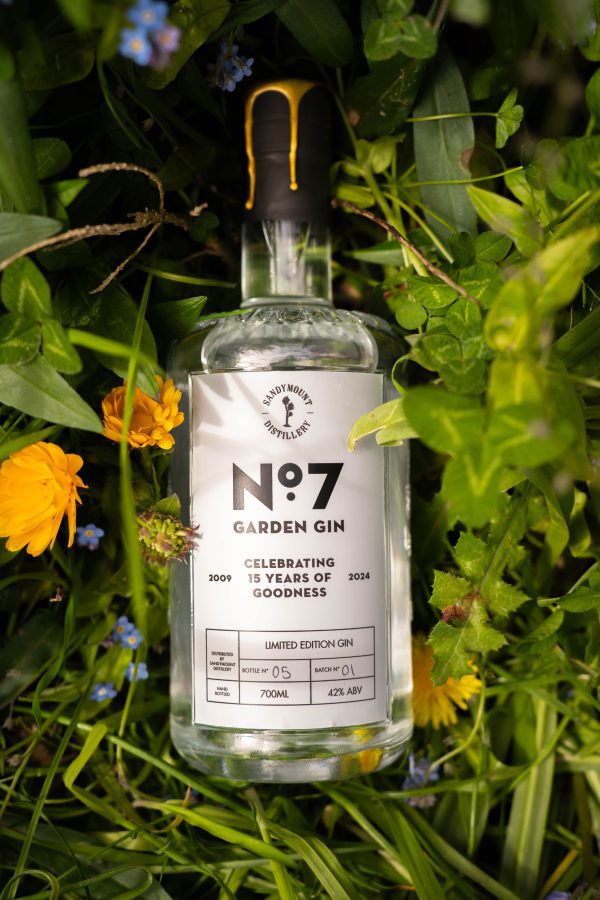 A clear glass bottle of gin with a label on the front titled "No.7 Garden Gin". The bottle is placed in a field pf green grasses and flowers.